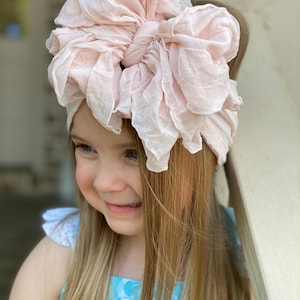 May include: A young girl with long blonde hair is wearing a light pink ruffled headband. The headband is tied in a large bow on top of her head.