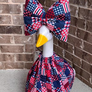 May include: A white goose figurine wearing a red, white, and blue patchwork fabric outfit. The ensemble includes a large, structured bow headpiece and a gathered, ruffled dress, both featuring a mix of plaid, gingham, and star-patterned fabric.
