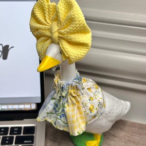 May include: A white ceramic goose figurine wearing a bright yellow textured fabric bow on its head and a ruffled collar featuring a mix of yellow gingham, blue floral, and yellow floral patterns. The goose has a yellow beak and yellow feet.