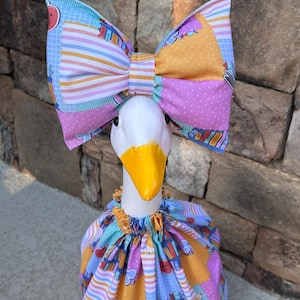 May include: A white goose figurine wearing a fabric outfit featuring a colorful pattern of books, pencils, and apples. The goose has a bright yellow beak and wears a large, matching fabric bow on its head, with a gathered, ruffled collar at the neck.