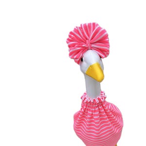 May include: A white goose wearing a pink and white striped turban and matching dress. The goose has a yellow beak and is set against a white background. The dress has a ruffled neckline.