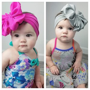 May include: Two babies wearing headbands with large bows. The headband on the left is pink and the headband on the right is gray. The baby on the left is wearing a floral swimsuit and the baby on the right is wearing a gray and white dress with colorful designs.