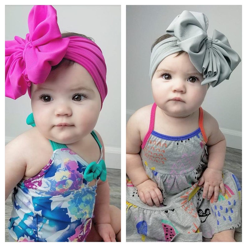May include: Two babies wearing headbands with large bows. The headband on the left is pink and the headband on the right is gray. The baby on the left is wearing a floral swimsuit and the baby on the right is wearing a gray and white dress with colorful designs.