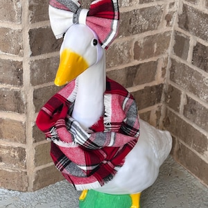 May include: A white ceramic goose figurine featuring a yellow beak and feet, wearing a red, white, and black plaid fabric infinity scarf around its neck and a matching plaid fabric bow attached to a white headband on its head.