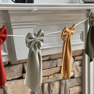 Può includere: Una ghirlanda decorativa con fiocchi di tessuto nei toni del rosso, grigio, oro e verde oliva. I fiocchi sono attaccati a uno spago bianco e appesi davanti a un camino in pietra. I fiocchi hanno un aspetto strutturato.