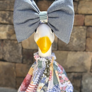May include: A decorative goose figurine with a large denim blue bow and a patchwork dress. The goose has a white head, a yellow beak, and black eyes. The dress is made of various patterned fabrics in shades of red, blue, and white.