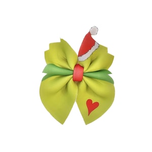 May include: Green and red Christmas bow with a Grinch-themed design. The bow features a green bow with a red center and a red Santa hat on top. The bow also has a red heart on one side.