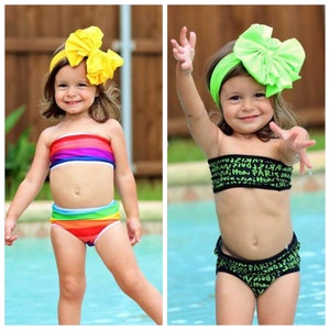 May include: Two young girls wearing different swimsuit styles. The girl on the left is wearing a rainbow striped bandeau top and matching bottoms. The girl on the right is wearing a black bandeau top with a green and black print and matching bottoms. Both girls are wearing large bows in their hair.