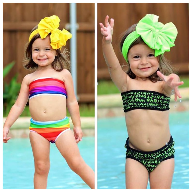 May include: Two young girls wearing different swimsuit styles. The girl on the left is wearing a rainbow striped bandeau top and matching bottoms. The girl on the right is wearing a black bandeau top with a green and black print and matching bottoms. Both girls are wearing large bows in their hair.