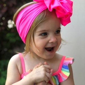 May include: A young girl wearing a bright pink headband with a large bow and a rainbow striped swimsuit. She is smiling and has her hands clasped together.