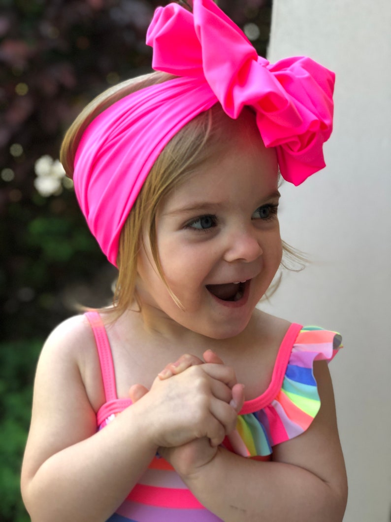 May include: A young girl wearing a bright pink headband with a large bow and a rainbow striped swimsuit. She is smiling and has her hands clasped together.