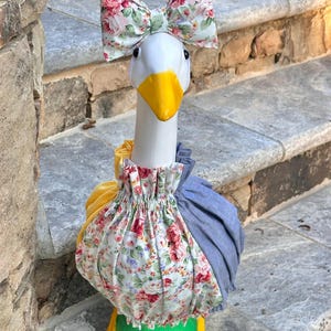 May include: A decorative goose statue with a white head, yellow beak and feet, and a floral bow. The goose wears a floral and blue fabric dress with a yellow accent. The statue is placed on a stone staircase.