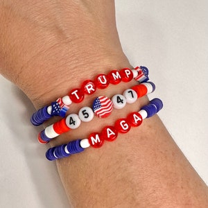 May include: Three beaded bracelets with red, white, and blue beads. The top bracelet spells out "TRUMP" in red beads. The middle bracelet has two American flag beads and the numbers "45" and "47" in white beads. The bottom bracelet spells out "MAGA" in red beads.