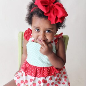May include: A young girl wearing a red and white strawberry print dress with a large red bow in her hair. She is sitting on a green chair and has her finger in her mouth.