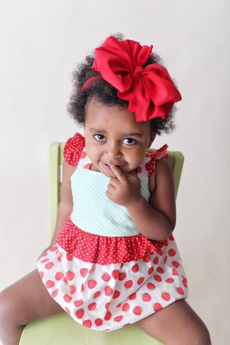 May include: A young girl wearing a red and white strawberry print dress with a large red bow in her hair. She is sitting on a green chair and has her finger in her mouth.