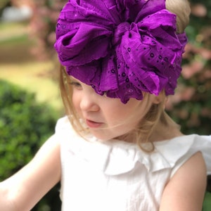 May include: A young girl wearing a white dress with a large purple bow headband with sequins. The headband is made of ruffled fabric.