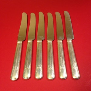 May include: Six stainless steel dinner knives with ornate handles are arranged on a red surface. The knives have a classic design, with a slightly curved blade and a decorative pattern on the handle. The word "STAINLESS" is visible on the blade.