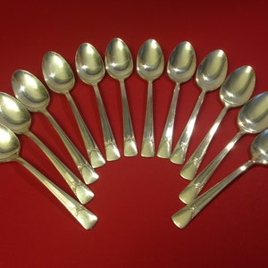 May include: A set of twelve silver spoons with ornate handles arranged in a semi-circle on a red background.
