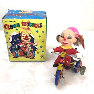 May include: A vintage mechanical clown tricycle toy with a box. The clown figure wears a red outfit and a pink hat. The tricycle is yellow and blue. The box is yellow with the words "Clown Tricycle" and a picture of a clown on a tricycle.