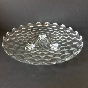 May include: A clear glass cake stand with a scalloped edge and a diamond pattern. The stand has three feet.