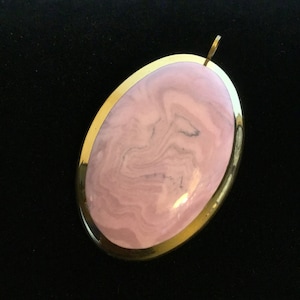 May include: A pink and white oval pendant with a gold rim. The pendant has a swirling pattern.