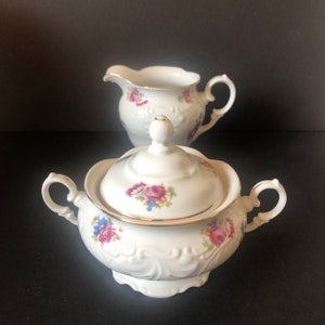 May include: White porcelain sugar bowl and creamer set with a floral design. The sugar bowl has a lid and both pieces have gold trim.