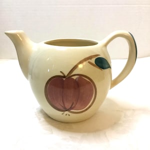 May include: Cream-colored ceramic pitcher with a rounded body, spout, and handle. The pitcher is decorated with a large red apple design with brown outlines and a green leaf. A charming addition to any kitchen.