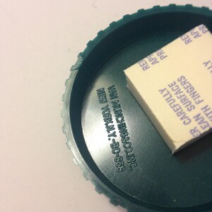 May include: A green plastic lid with a white adhesive sticker. The sticker has the text "Clean Surface Carefully With Fingers" and "Van Munching & Co. Inc. New York, N.Y. - BC-868".