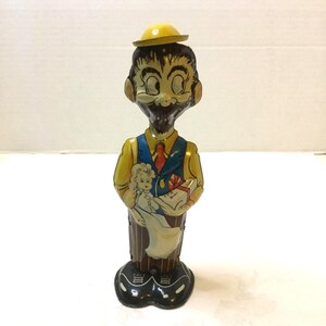 May include: Vintage tin wind-up toy of a man in a yellow hat, yellow shirt, blue vest, and red tie. The toy holds a doll and a gift box labeled "FOR BABY". The base is black with white accents. The toy has a painted face with a mustache and beard.