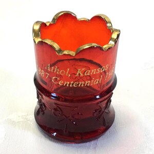 May include: A red glass cup with a gold rim and a gold inscription that reads "1887 Athol, Kansas Centennial 1987".