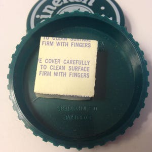 May include: Green plastic bottle cap with a white sticker that says "To clean surface firm with fingers" and "Ve cover carefully to clean surface firm with fingers". The cap also has the word "Heineken" printed on it in white letters.