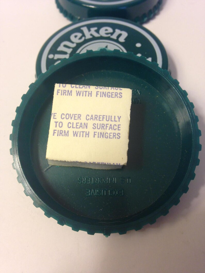 May include: Green plastic bottle cap with a white sticker that says "To clean surface firm with fingers" and "Ve cover carefully to clean surface firm with fingers". The cap also has the word "Heineken" printed on it in white letters.