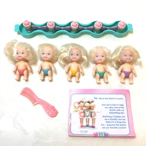 May include: A set of five baby dolls with blonde hair and blue eyes, each wearing a different colored diaper. Accessories include a pink comb, a bottle rack with five bottles, and a booklet that reads "Hi! We're the Quints Cousins!"