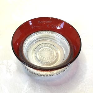 May include: A clear glass bowl with a red rim and a beaded edge. The bowl has a decorative pattern on the bottom and says "Souvenir Forman N. Dakota" on the rim.