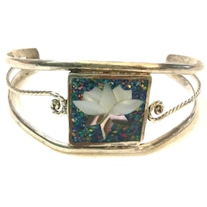 May include: Silver cuff bracelet with a square centerpiece featuring a white lotus flower inlay. The square is surrounded by a mosaic of colorful stones. The bracelet has a twisted rope design and a floral accent on each side.