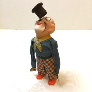 May include: Vintage wind-up toy clown with a black top hat balanced on its nose. The clown wears a blue shirt, plaid pants, and red shoes. A yellow collar and a small badge are also visible. The toy is in a standing position.