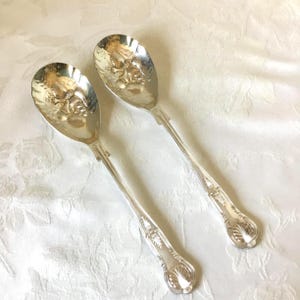 May include: Two antique silver serving spoons. The spoons have an oval bowl with a detailed embossed design and long, slender handles. The handles feature ornate detailing and end in a shell-shaped design. The spoons are placed on a white, textured surface.