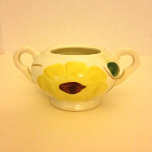 May include: A small, cream-colored ceramic sugar bowl with two handles. The bowl is decorated with a large yellow flower with a dark brown center. Green leaves and stems accent the flower design. The bowl sits on a small, round base.