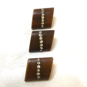 May include: Three brown, diamond-shaped buttons with a vertical row of small, clear rhinestones. The buttons have a glossy finish and are set against a white background. These buttons are suitable for sewing projects or adding embellishments to clothing.