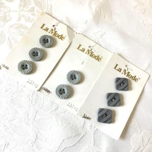 May include: Three cards of La Mode brand buttons. The cards feature gray buttons in two styles: round with a ridged edge and square with diagonal stripes. Each button has a black center. The cards are on a white lace surface.