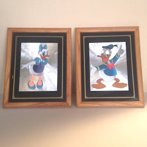 May include: Two framed pictures of Donald Duck and Daisy Duck. The pictures are in wooden frames with a black mat. The pictures are of the characters in a silver foil finish.
