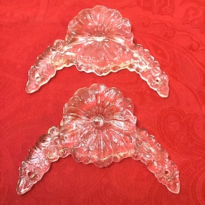 May include: Two clear glass decorative pieces with a floral design. Each piece has a central flower shape and curved, ornate arms. The glass has a slight pink hue, and the pieces are set against a red background.