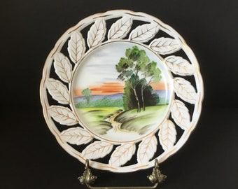 Puede incluir: Plato de porcelana blanca con un diseño de hojas doradas y una escena de paisaje pintada. El plato está en un soporte de metal.