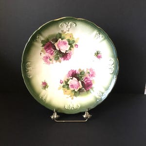 May include: A large, white porcelain plate with a green rim and a floral design featuring pink roses. The plate is on a metal stand.