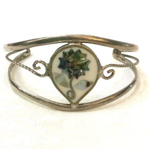 May include: Silver-toned cuff bracelet with a central teardrop-shaped design. The design features a flower made of iridescent abalone shell pieces set against a white background. The bracelet has a double-band design with decorative swirls.