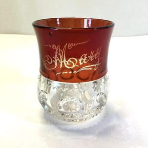 May include: A clear glass goblet with a red glass rim and gold lettering that says "Mary". The goblet has a textured pattern of stars and circles.