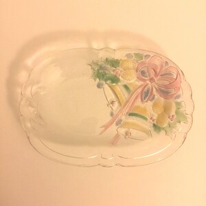 May include: Clear glass serving platter with a scalloped edge. The platter features a painted design of Christmas bells, pinecones, and a pink bow. The bells are yellow and green, and the pinecones are yellow.