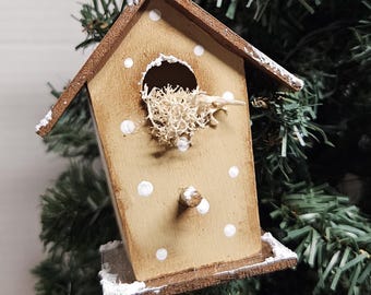 May include: A wooden birdhouse ornament with a brown roof and white polka dots. The birdhouse is filled with dried moss and is hanging from a twine string on a green Christmas tree.