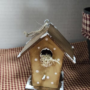 K&ouml;nnte beinhalten: Ein kleines Vogelhaus aus Holz mit braunem Dach und W&auml;nden, verziert mit wei&szlig;en Punkten und Kunstschnee. Das Vogelhaus hat eine runde &Ouml;ffnung, die mit getrocknetem Moos gef&uuml;llt ist. Ein rustikales Deko-Objekt.