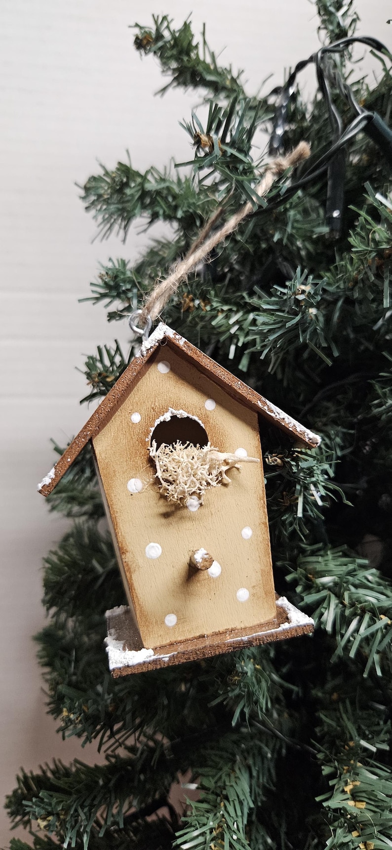 K&ouml;nnte beinhalten: Ein Holz-Vogelhaus-Ornament mit braunem Dach und wei&szlig;en Tupfen. Das Vogelhaus ist mit getrocknetem Moos gef&uuml;llt und h&auml;ngt an einer Schnur an einem gr&uuml;nen Weihnachtsbaum.